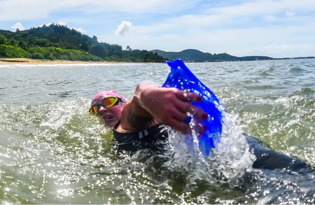 Fotografia de uma nadadora no mar, usando touca rosa e óculos, enquanto segura um saco plástico azul durante a braçada. Ao fundo, faixa de areia, vegetação e morros sob céu claro.”