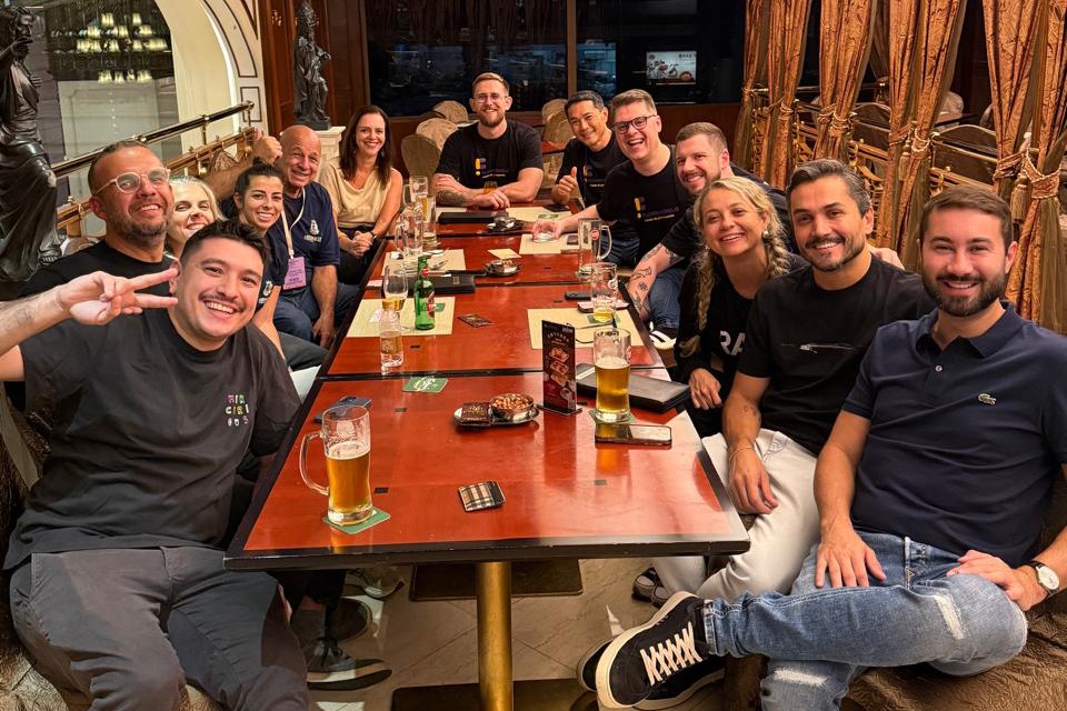 Fotografia de grupo em restaurante elegante: doze adultos sorriem ao redor de uma mesa longa com copos de cerveja, e o homem em primeiro plano faz sinal de paz para a câmera. 