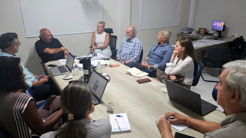 Fotografia de reunião em escritório: nove pessoas sentadas ao redor de uma mesa, ouvindo um homem que fala e gesticula. Sobre a mesa, há laptops, cadernos, xícaras e garrafa térmica. Ao fundo, mesa auxiliar com papéis e monitor ligado.” 