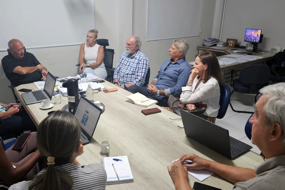 Fotografia de reunião em escritório: nove pessoas sentadas ao redor de uma mesa, ouvindo um homem que fala e gesticula. Sobre a mesa, há laptops, cadernos, xícaras e garrafa térmica. Ao fundo, mesa auxiliar com papéis e monitor ligado.