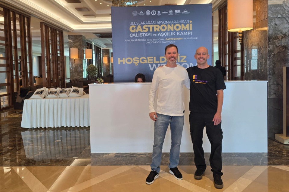 Fotografia de dois homens sorridentes em lobby de hotel, diante de painel de workshop internacional de gastronomia.