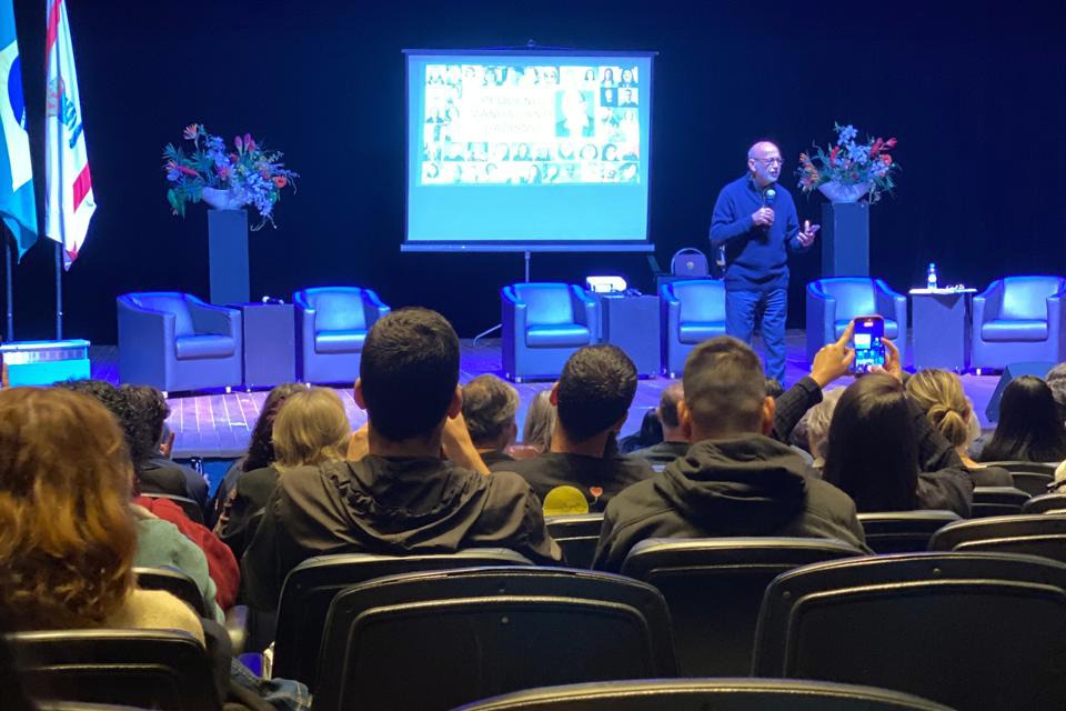 Fotografia de palestra em auditório, com público sentado diante de um homem falando ao microfone em um palco iluminado.