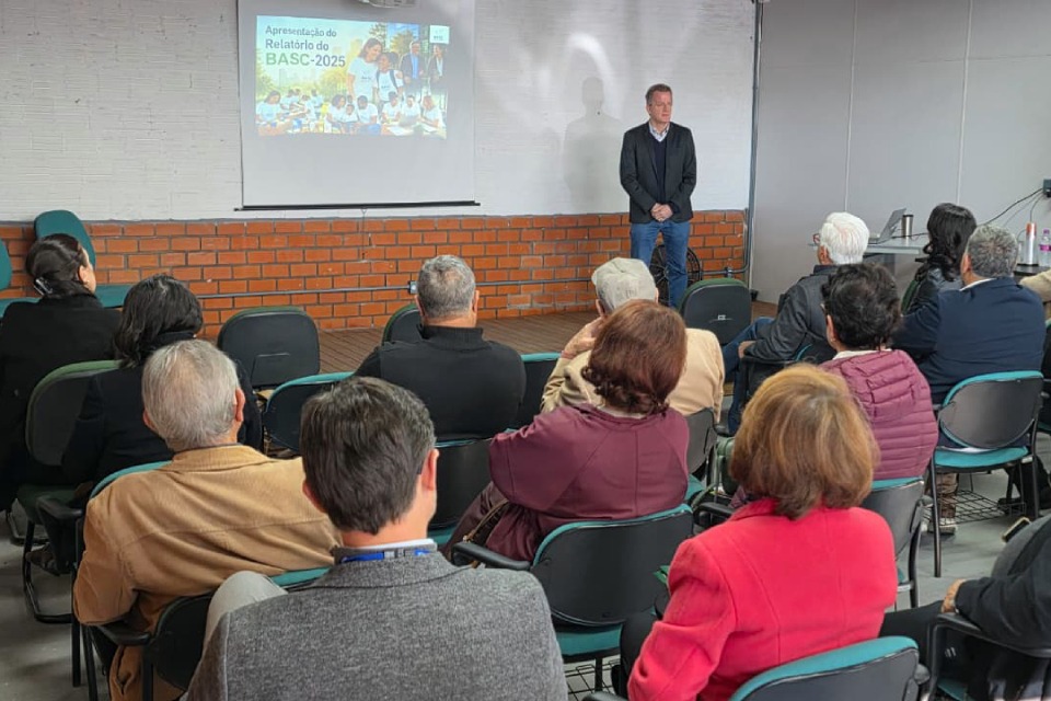 Fotografia de apresentação em sala, com público sentado diante de um homem e projeção do Relatório do BASC.
