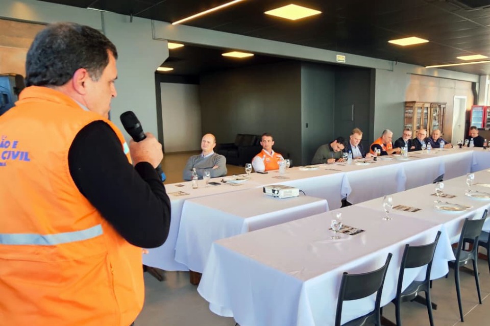 Fotografia de reunião com representante da Defesa Civil falando ao microfone para participantes sentados em mesas.