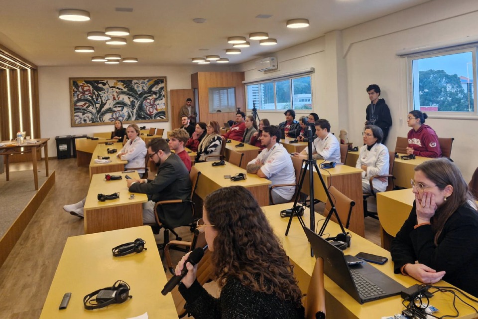 Fotografia de reunião em sala com mesas amarelas, onde profissionais acompanham uma apresentação e uma mulher fala ao microfone.