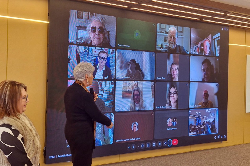 Fotografia de reunião híbrida, com duas mulheres diante de um telão exibindo participantes em videoconferência.