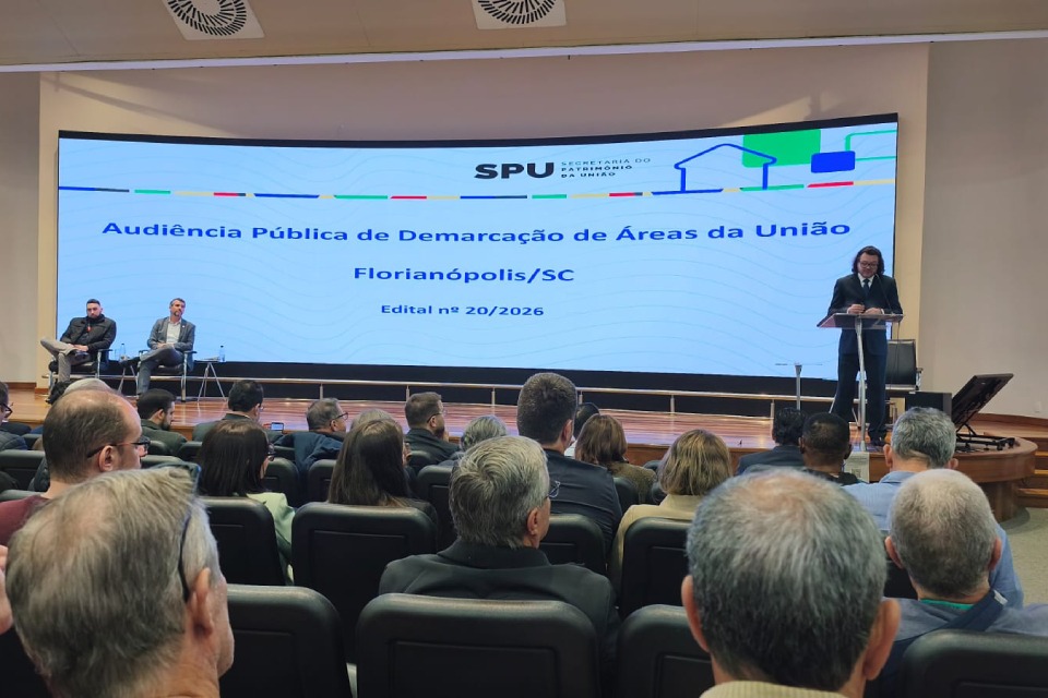 Fotografia de audiência pública em auditório, com palestrante ao púlpito e telão sobre demarcação de áreas da União.