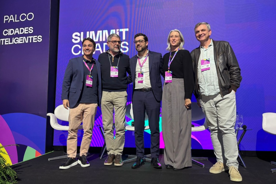 Fotografia de cinco participantes no palco do Summit Cidades, em frente a painel roxo sobre cidades inteligentes. 