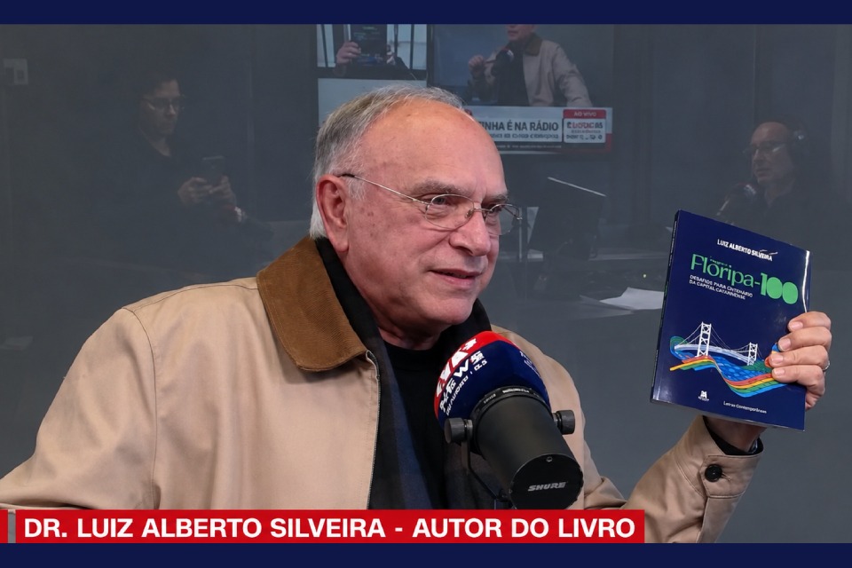 Fotografia de entrevista em rádio, com Luiz Alberto Silveira segurando o livro “Floripa mais cem” diante de um microfone.