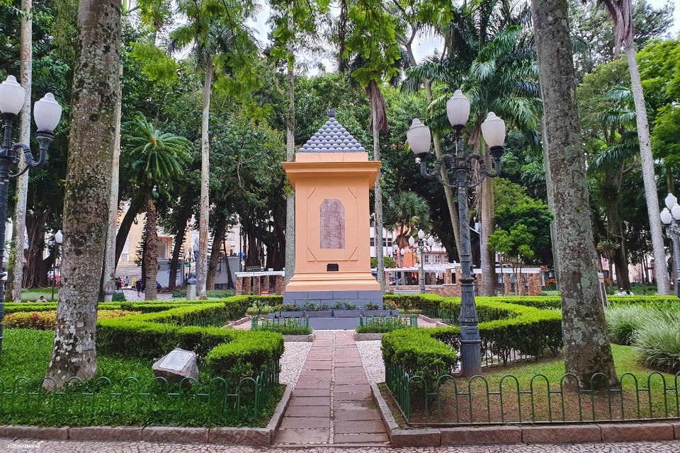 Fotografia de praça arborizada, com monumento central, caminhos ajardinados, postes de luz e vegetação densa.