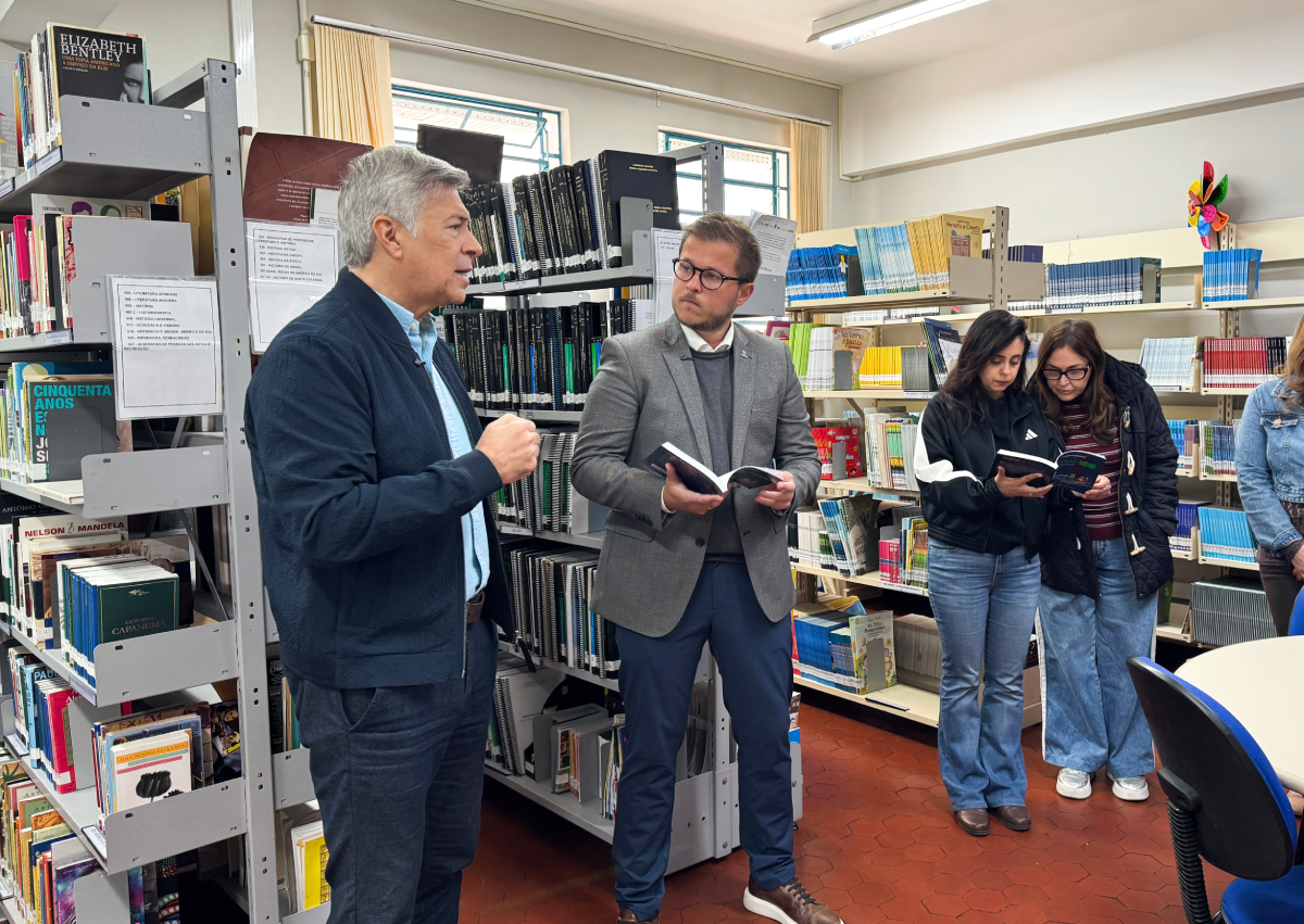 Fotografia mostra um homem falando em uma biblioteca enquanto outro segura um livro aberto; ao lado, duas mulheres leem juntas entre as estantes.