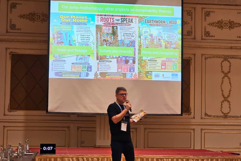 Fotografia de homem com microfone e material impresso durante palestra; ao fundo, projeção sobre sustentabilidade com os temas “Our Planet, Our Home”, “Roots That Speak” e “Earthworm in Head”.