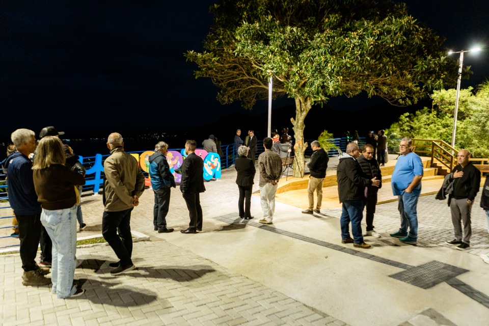 Fotografia noturna mostra cerca de vinte pessoas conversando em um mirante iluminado, junto à lagoa, com árvore e montanhas ao fundo.