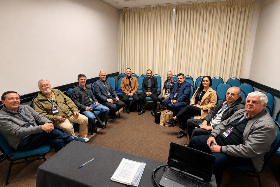 Fotografia mostra dez homens e uma mulher sentados em círculo em uma sala de reunião, todos usando crachás de evento.