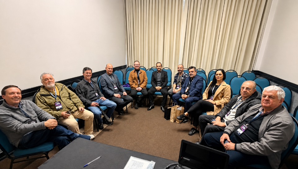 Fotografia mostra dez homens e uma mulher sentados em círculo em uma sala de reunião, todos usando crachás de evento. 