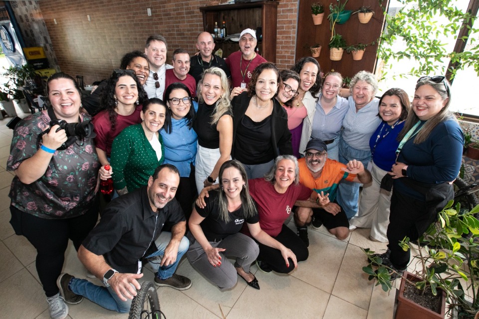 Fotografia mostra cerca de vinte pessoas sorridentes reunidas em grupo em um ambiente interno com plantas; algumas estão agachadas à frente.