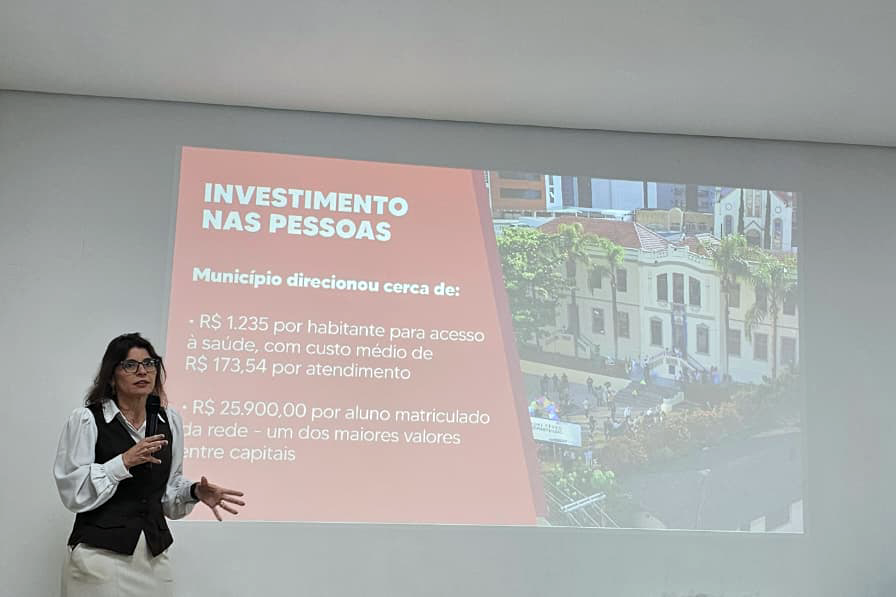 Fotografia de uma mulher apresentando um slide. Texto: “Investimento nas pessoas. Município direcionou cerca de: mil duzentos e trinta e cinco reais por habitante para acesso à saúde, com custo médio de cento e setenta e três reais e cinquenta e quatro centavos por atendimento; vinte e cinco mil e novecentos reais por aluno matriculado na rede — um dos maiores valores entre capitais”.