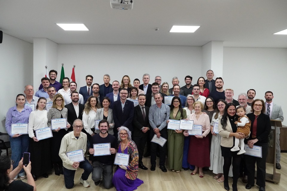 Fotografia de dezenas de pessoas reunidas em uma sala, várias segurando certificados de posse e posando para a câmera.