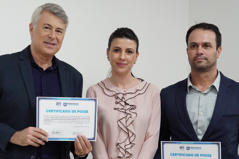 Fotografia de dois homens e uma mulher posando lado a lado; os homens seguram certificados de posse da Prefeitura de Florianópolis.