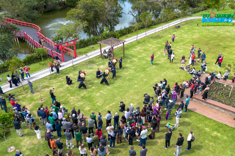 Fotografia aérea de uma apresentação de tambores no gramado do Jardim Japonês. O público acompanha a cena; ao fundo, uma ponte vermelha cruza o lago. No canto superior direito, lê-se “Eu amo o Botânico de Florianópolis.