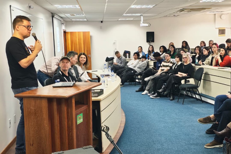 Fotografia de homem falando ao microfone diante de uma plateia sentada em sala de aula, com três pessoas à mesa.