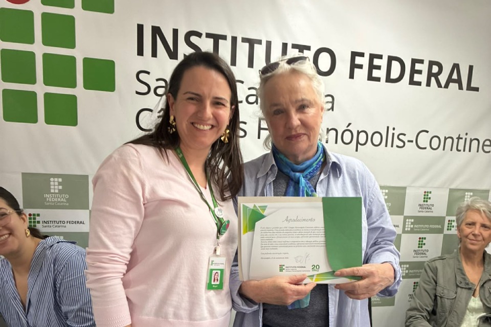 Duas mulheres posam lado a lado, uma delas segurando um certificado com o título “Agradecimento”, diante de um painel do Instituto Federal de Santa Catarina.
