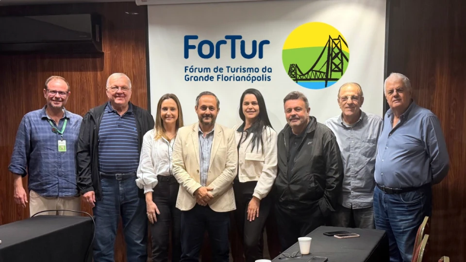 Fotografia de oito pessoas em pé, lado a lado, durante encontro do ForTur, diante do painel “ForTur – Fórum de Turismo da Grande Florianópolis”.