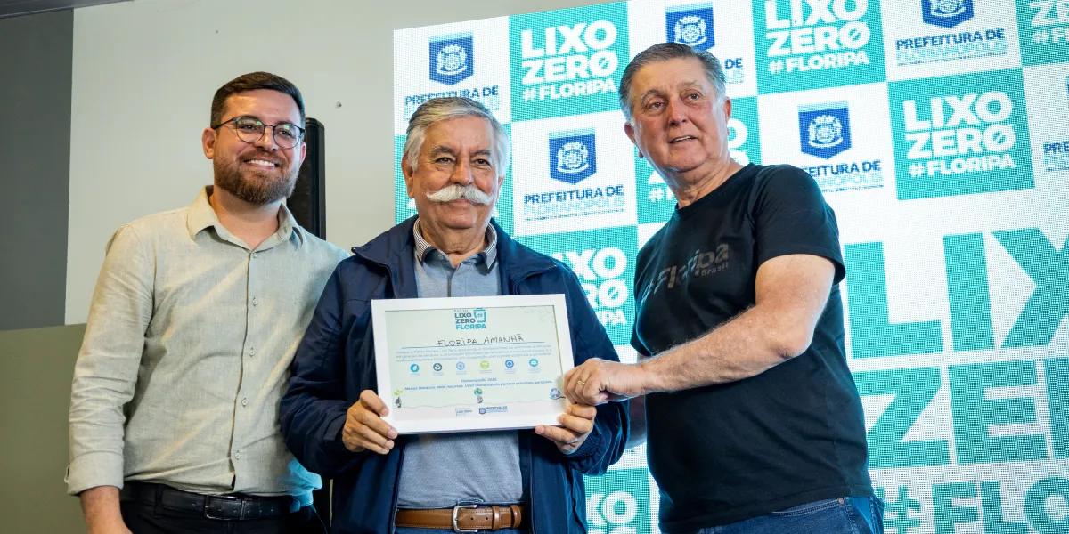 Três homens posam diante de painel do Lixo Zero Floripa. Ao centro, um deles segura um certificado emoldurado.