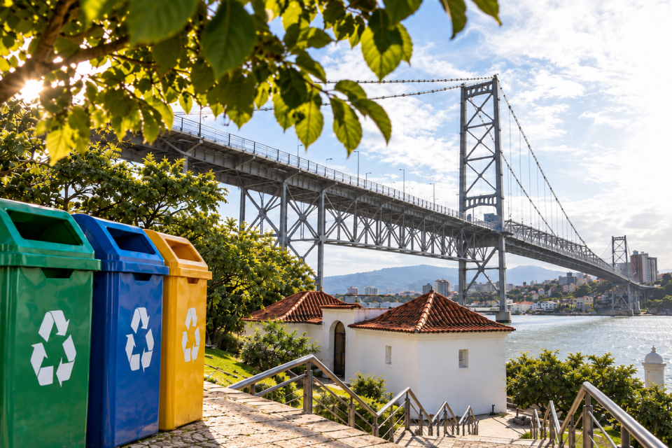 Fotografia de três lixeiras de reciclagem, nas cores verde, azul e amarela, à margem da água, com a Ponte Hercílio Luz ao fundo.