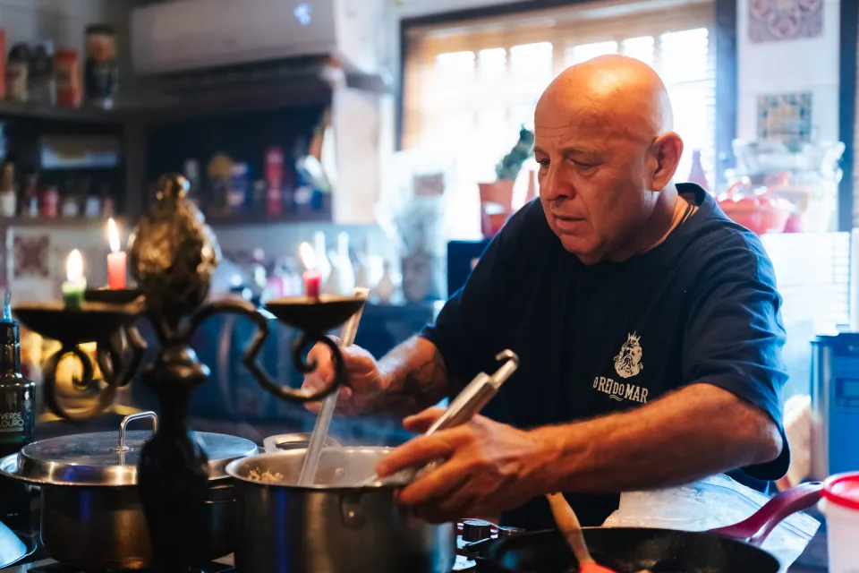 Fotografia de homem careca, com camiseta azul, mexendo uma panela no fogão de uma cozinha.