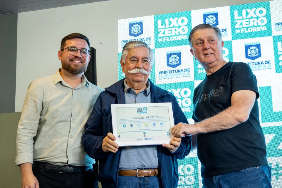 Três homens posam diante de painel do Lixo Zero Floripa. Ao centro, um deles segura um certificado emoldurado.