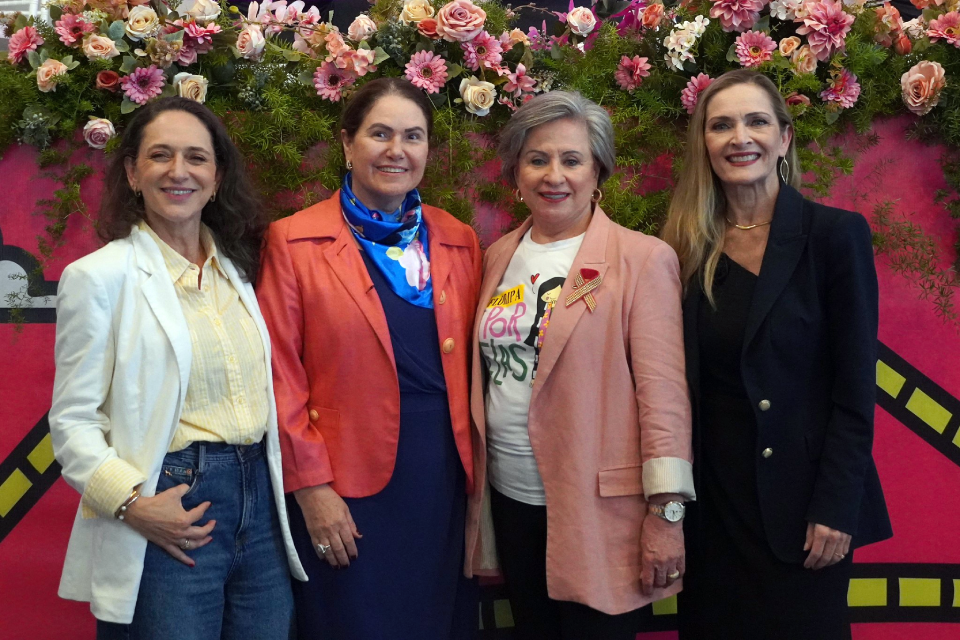 Fotografia de quatro mulheres sorridentes lado a lado, diante de um painel decorado com flores em tons de rosa, branco e creme.